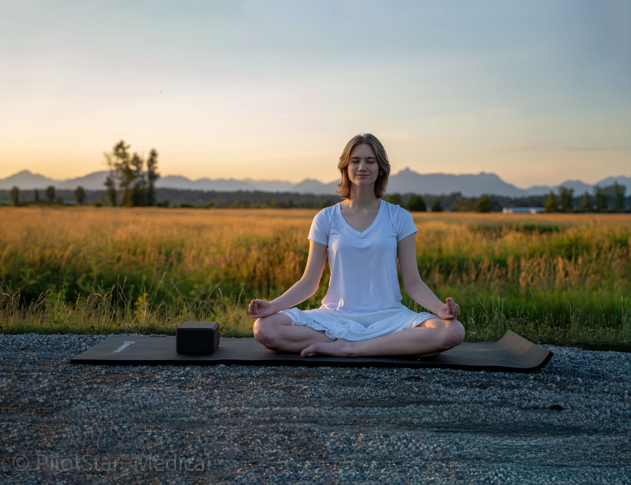 Woman meditating on a yoga mat beside a field at sunset